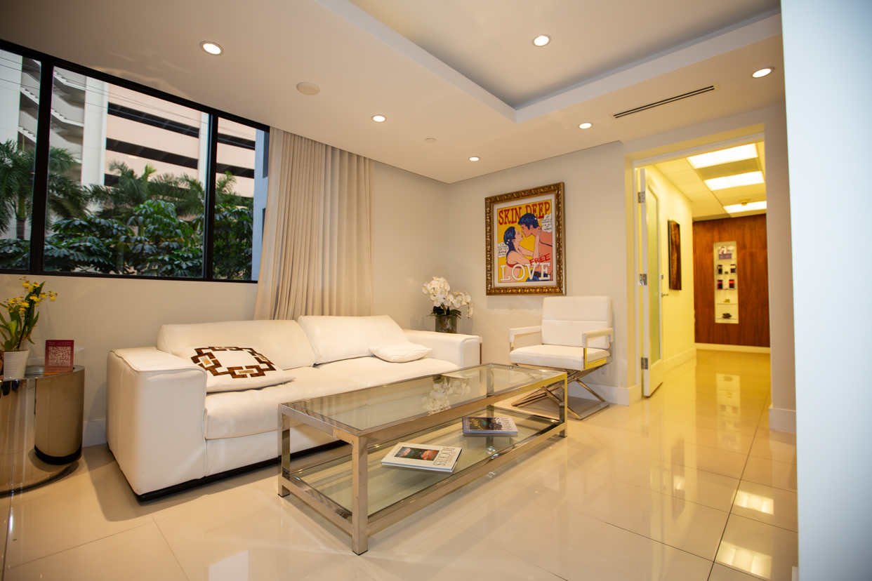 Dr. Alan Serure’s plastic surgery practice patient sitting area with white sofa, marble floor, glass coffee table, and colorful painting.